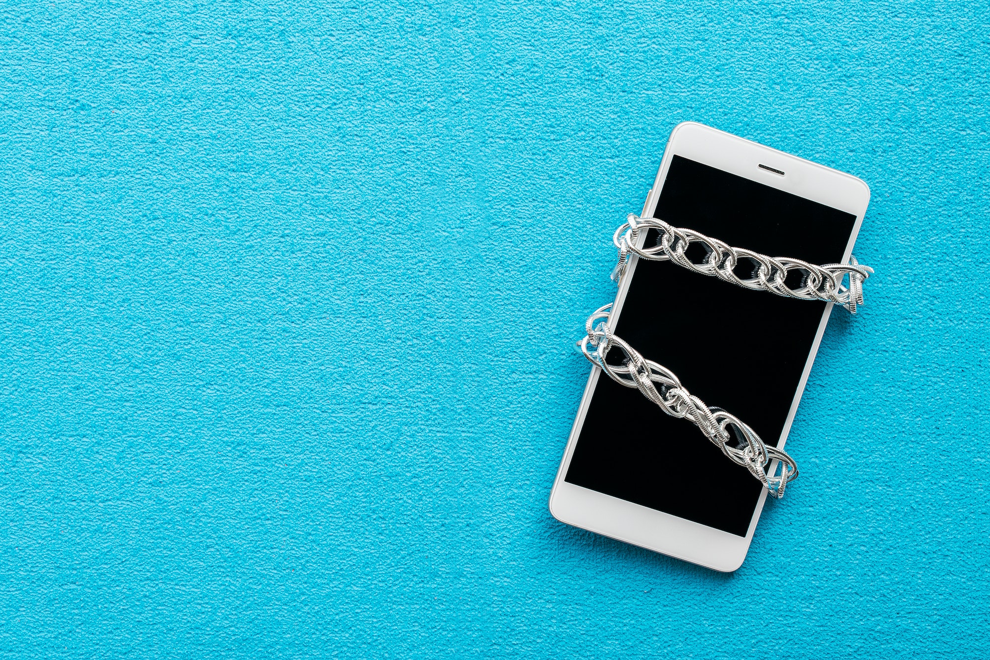 White smartphone with metal chain on blue background. Digital detox, dependency on tech, no gadget