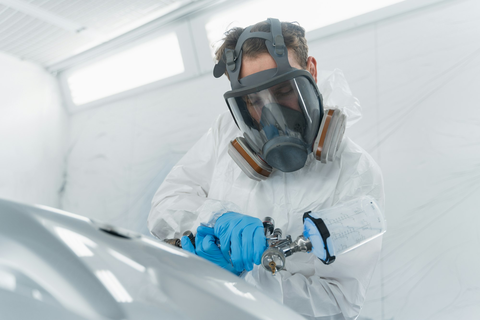 Auto service worker painting car element with spray gun in a paint chamber.