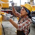 Young Hispanic woman in coveralls and safety helmet looking at working plan